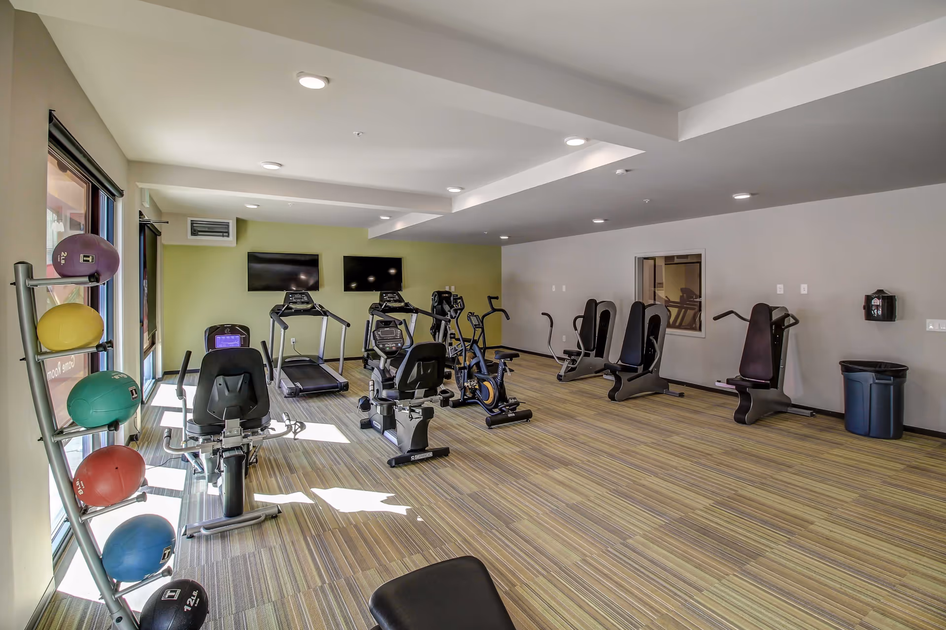 Bright interior fitness room with treadmills, stationary bikes, weight machines and a rack of colorful medicine balls.