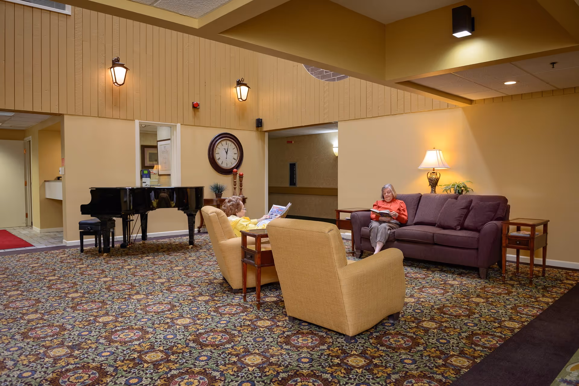 A cozy common area in a retirement center featuring two elderly women seated and reading. One woman is sitting on a purple couch near a side table with a lamp and plant, while the other is seated in a yellow armchair. The room has a patterned carpet, a black grand piano, a large wall clock, and warm yellow walls with wall-mounted lights.