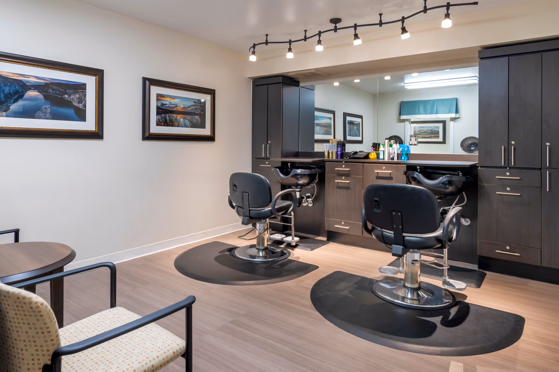 Interior view of a salon area with two black salon chairs in front of a large mirror and dark wood cabinetry. The room has light-colored walls with two framed landscape photographs and a small seating area with a round table and two chairs.