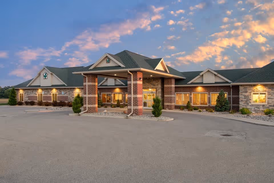 Exterior view of a single-story senior living facility building with a covered entrance supported by brick columns, surrounded by small shrubs and trees, under a partly cloudy sky at sunset.