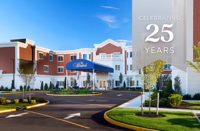Front entrance of The Bristal assisted living building with a blue canopy, landscaped driveway, and a 'Celebrating 25 Years' banner.