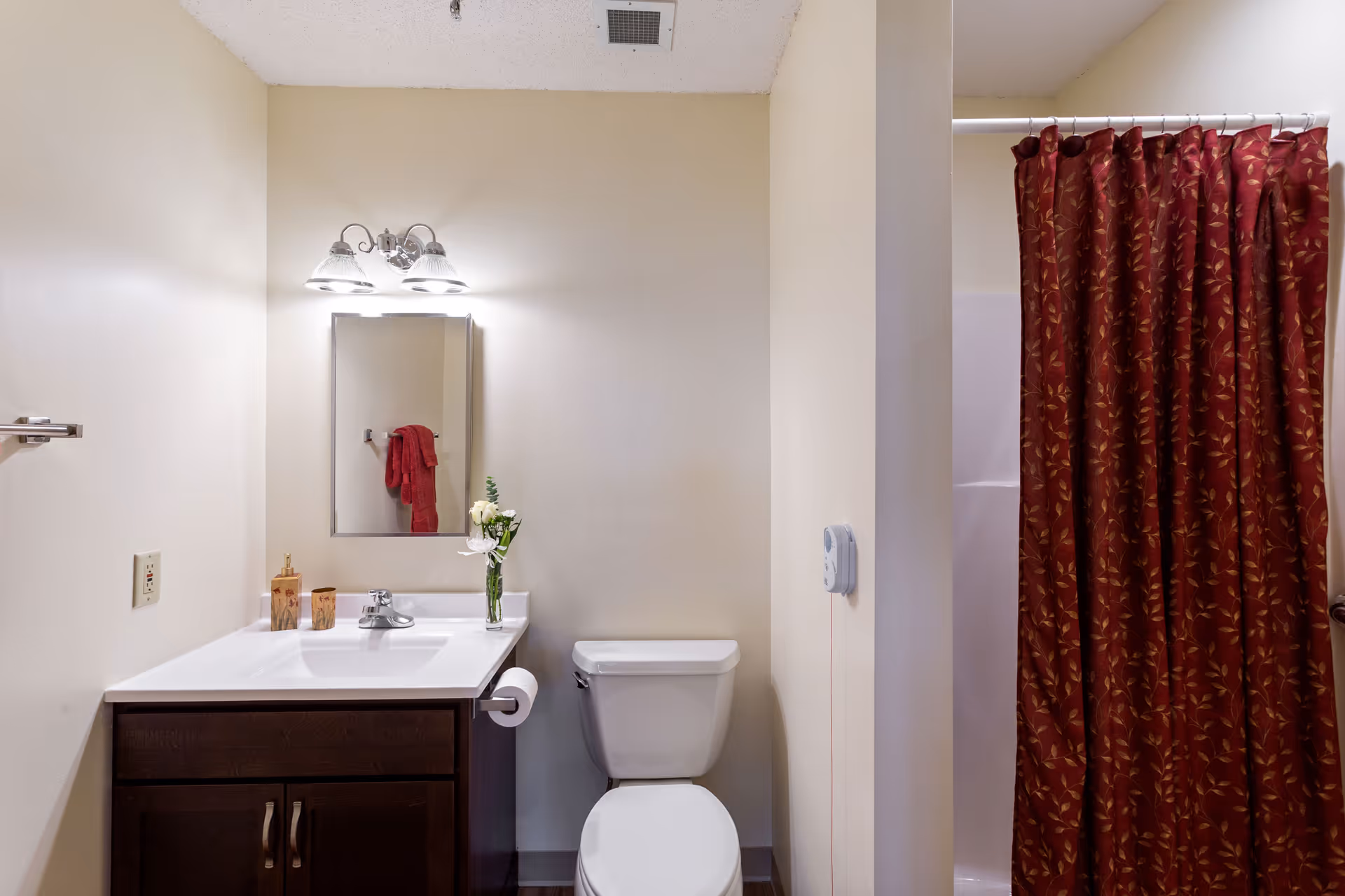 A clean bathroom with a white toilet and a dark wood vanity with a white countertop. Above the vanity is a mirror with three light fixtures. A vase with flowers and two small containers are on the countertop. To the right, there is a shower with a red patterned curtain.