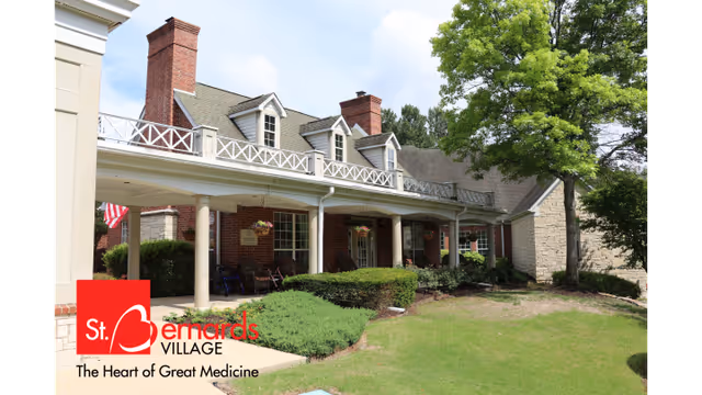 Exterior view of St. Bernards Village building with a covered porch, brick chimneys, and a well-maintained lawn with shrubs and a tree under a partly cloudy sky.