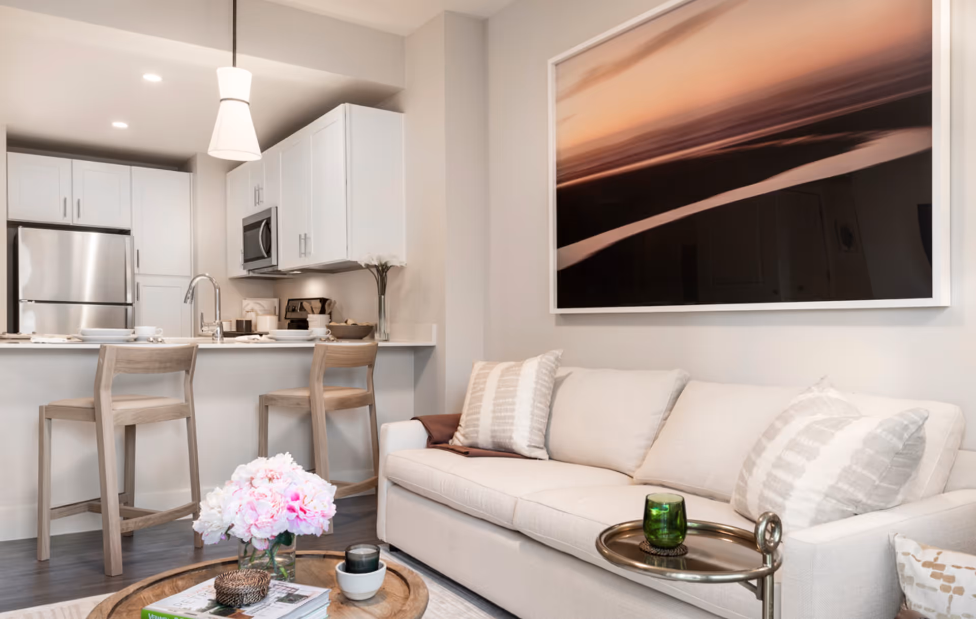 A bright open living room with a beige sofa, coffee table with flowers, bar stools at a kitchen island, and large framed artwork on the wall.