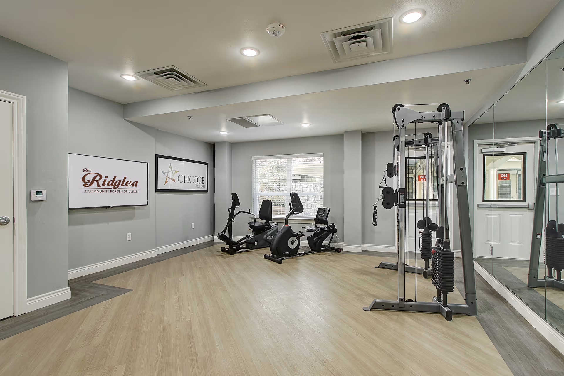 A fitness room in The Ridglea Senior Living facility featuring exercise equipment including two recumbent bikes and a multi-functional weight machine. The room has light wood flooring, gray walls, recessed ceiling lights, a large mirror on one wall, and a window letting in natural light. Two framed signs are mounted on the wall, one displaying 'The Ridglea A Community for Senior Living' and the other 'Choice Rehab'.