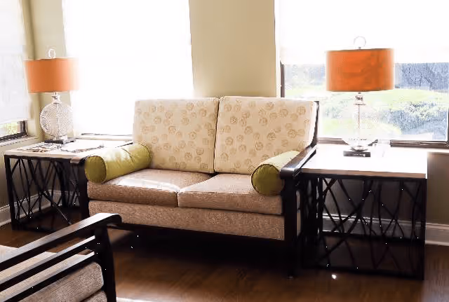 A cozy living room area with a beige patterned loveseat featuring green bolster pillows, flanked by two side tables with black lattice bases and white tops. Each table holds a lamp with a clear base and an orange lampshade. Large windows behind the loveseat let in natural light.