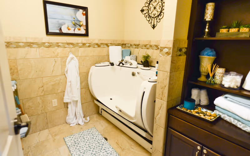 A bathroom with beige tiled walls and floor featuring a white walk-in bathtub with built-in controls. A white bathrobe hangs on the wall, and a patterned bath mat is on the floor. To the right, there is a dark wooden shelving unit holding folded towels, decorative items, and bath supplies. A framed picture and a decorative wall piece are mounted on the walls.