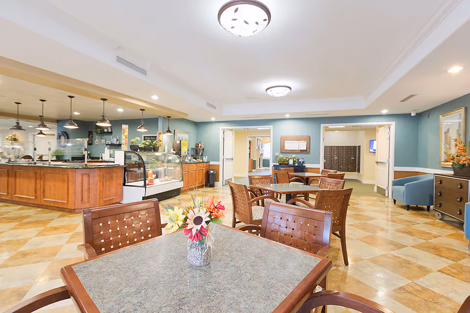 A bright and spacious dining area in a senior living facility with several wooden tables and chairs. The room features a checkered tile floor, blue-green walls, and ceiling lights. In the background, there is a counter with hanging pendant lights and a display case, likely for food service. There are also some decorative flowers on the tables and a seating area with blue chairs and a wooden cabinet on the right side.