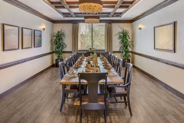 A long wooden dining table set for a meal with plates, napkins, and glasses in a well-lit dining room. The room features a coffered ceiling with two chandeliers, framed artwork on the walls, two tall green plants near a window with curtains, and wooden flooring.