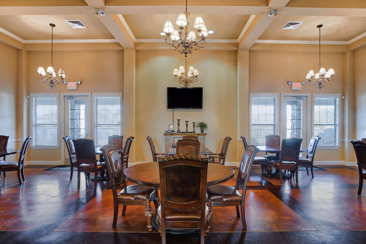 A dining room with multiple round wooden tables and matching chairs arranged neatly. The room features beige walls, three chandeliers with lampshades hanging from the ceiling, and a flat-screen TV mounted on the wall above a small cabinet with decorative items. There are windows with blinds and exit doors on either side of the TV.