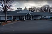 Front exterior of a single-story building with a covered entrance, driveway, and leafless trees under an overcast sky.