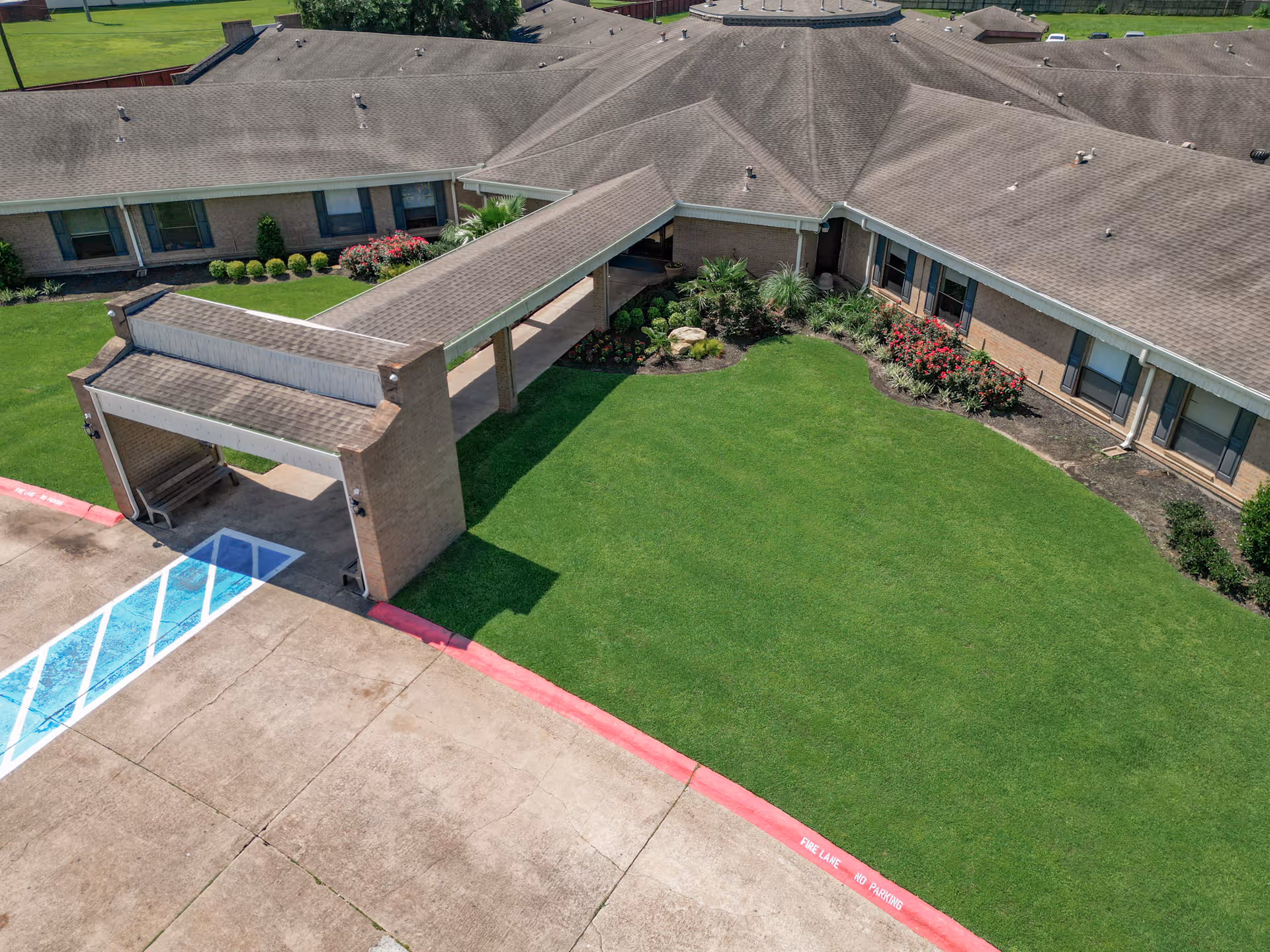 Aerial view of the exterior of a single-story nursing and rehabilitation facility with a covered entrance, well-maintained green lawn, flower beds, and a driveway with a marked handicap parking space.