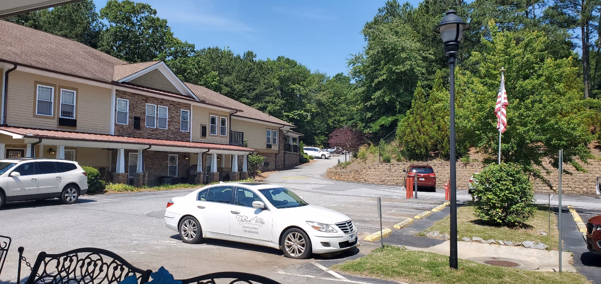 Front exterior of a two-story assisted living building with a parking lot, parked cars, a flagpole, and surrounding trees.