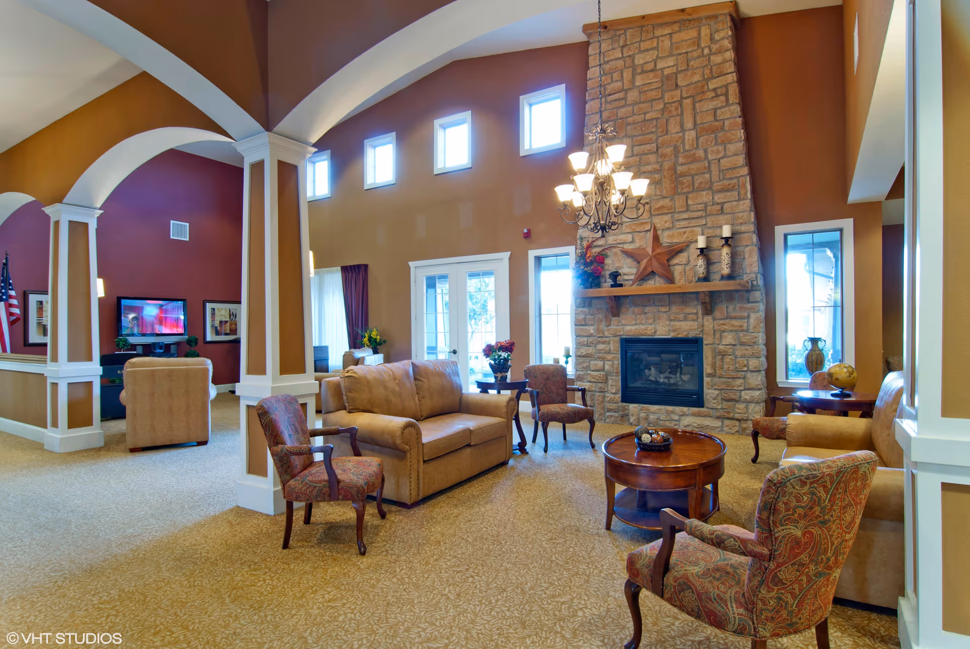 Bright assisted-living common room with vaulted ceilings, a stone fireplace, sofas, armchairs and a chandelier.