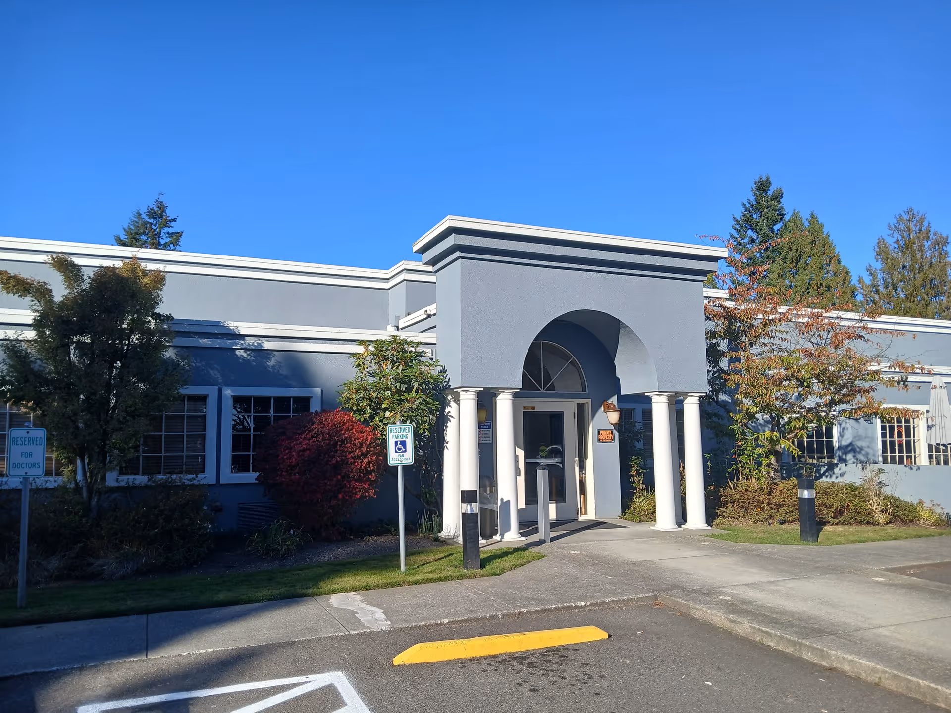 Exterior front view of a single-story building with a gray facade and white columns at the entrance. There are trees and bushes around the building, a clear blue sky, and parking spaces including a reserved handicapped parking spot near the entrance.