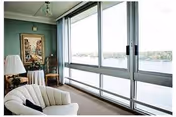 A bright living room with large floor-to-ceiling windows overlooking a body of water. The room features a white upholstered chair, a small round table with a lamp, a framed painting on the wall, and a decorative statue near the window.