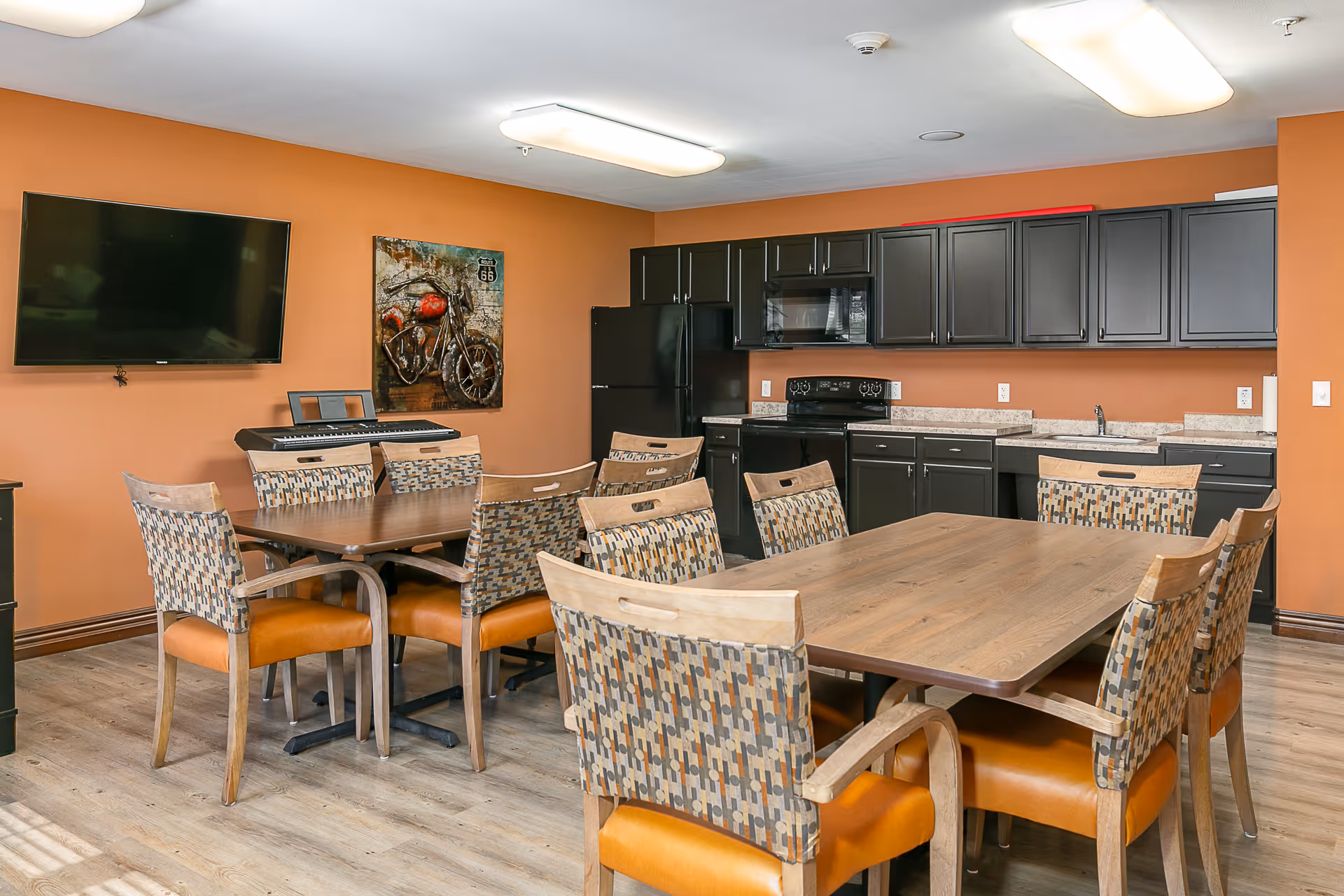 A communal dining area with two wooden tables surrounded by patterned chairs with orange cushions. The room features an orange wall with a mounted flat-screen TV and a painting of a motorcycle. There is a kitchenette with black cabinets, a black refrigerator, microwave, stove, and a sink along one wall. The floor is light-colored wood, and the ceiling has fluorescent lighting.