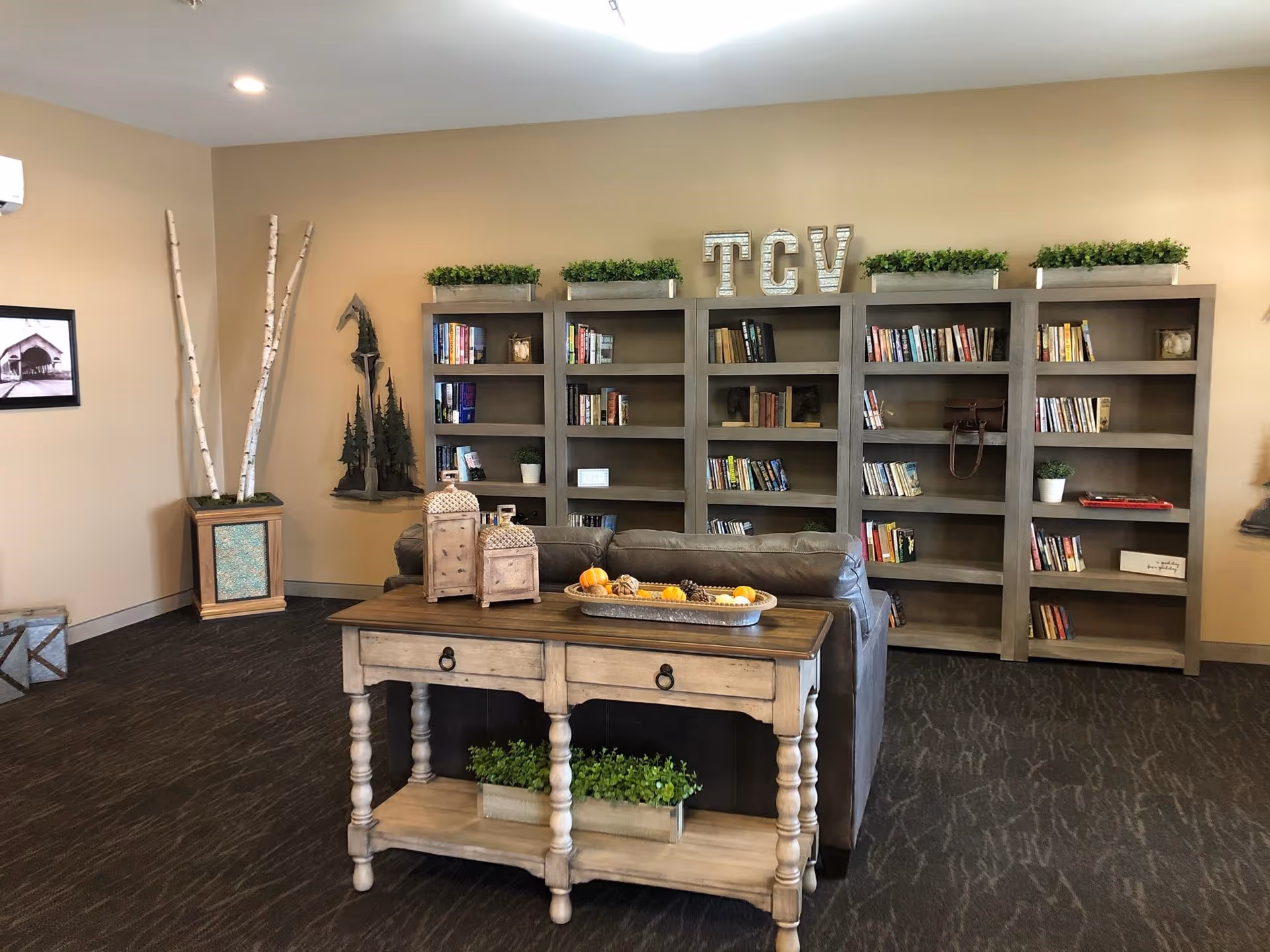 A cozy living room area with a brown leather couch facing a set of wooden bookshelves filled with books and decorative plants. On top of the bookshelves are large letters spelling 'TCV' and several green potted plants. In front of the couch is a wooden console table with decorative lanterns and a tray of seasonal decor. The room has beige walls, dark carpeted flooring, and additional decorative elements including tall birch branches in a planter and a framed picture on the wall.