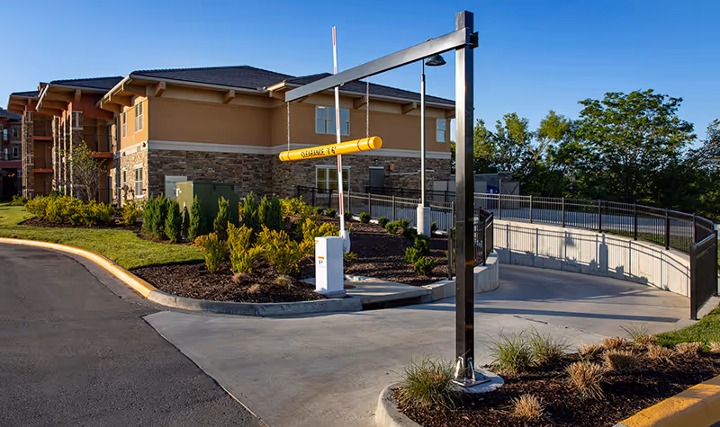 Exterior view of a senior living facility building with a stone and beige facade, surrounded by landscaped greenery and a paved driveway with a clearance bar and gate arm at the entrance.