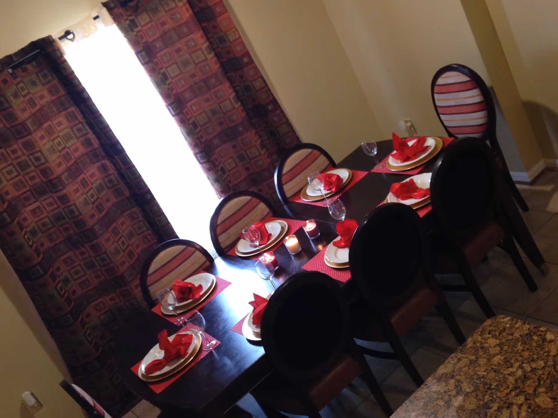 A dining room with a dark wooden table set for six people. Each place setting includes a white plate on a gold charger, a red folded napkin, and a wine glass. There are red placemats under each setting and three lit candles in the center of the table. The room has patterned curtains covering a window and beige walls.