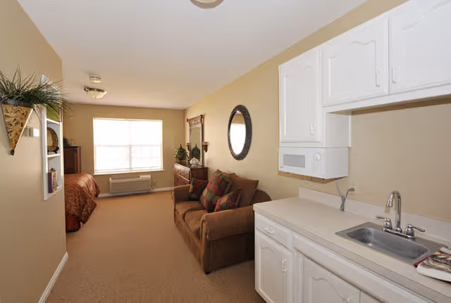 Interior view of a senior living facility room with a small kitchenette on the right, including a sink, microwave, and white cabinets. A brown couch with colorful pillows is positioned against the wall, and a round mirror hangs above it. In the background, there is a bed near a large window with blinds, a dresser, and some decorative plants and items.