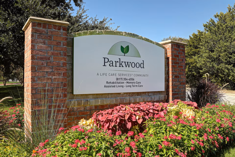 Outdoor sign for Parkwood, a life care services community, surrounded by colorful flowers and greenery under a clear blue sky.