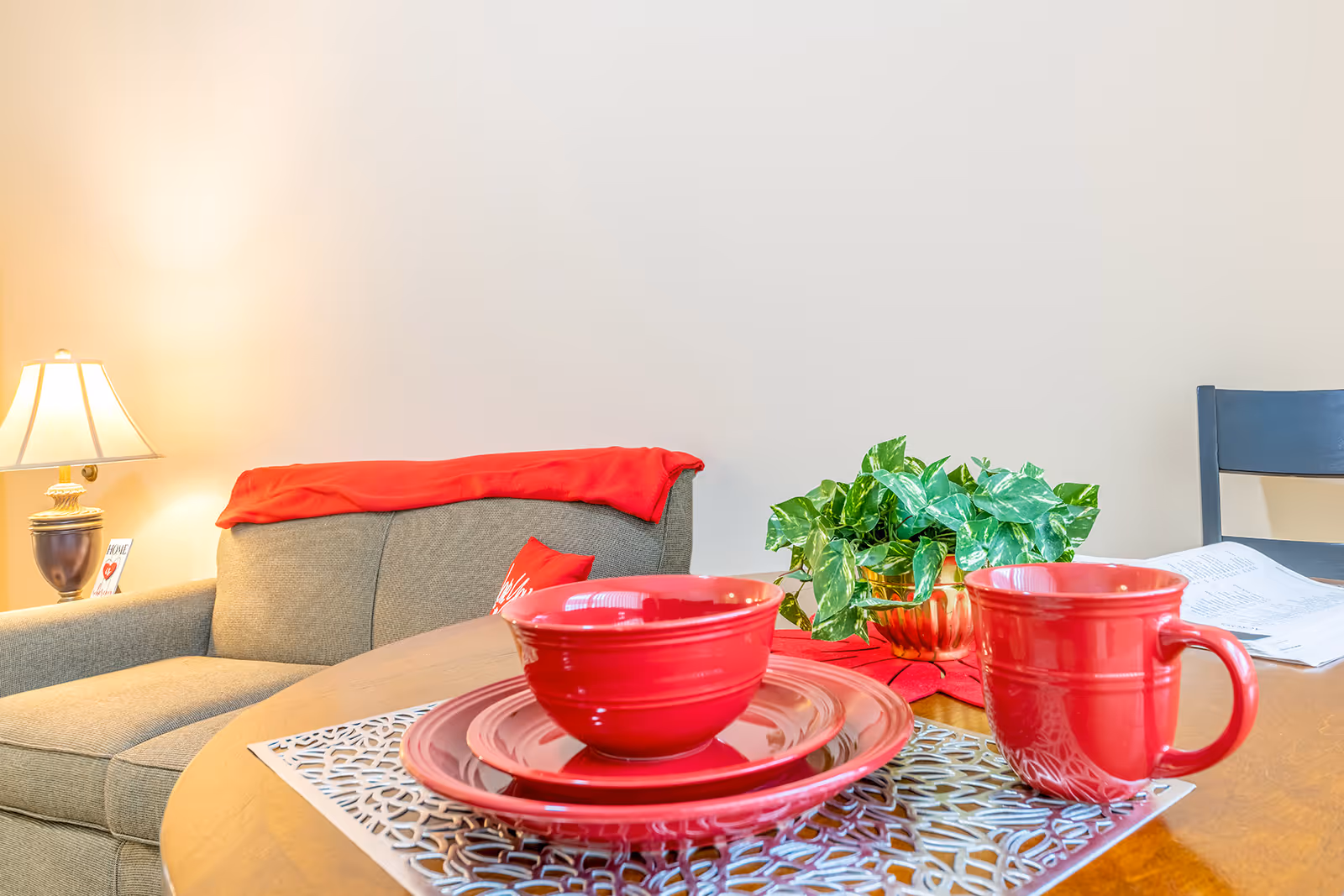 A cozy living area with a beige couch draped with a red blanket and a red pillow. In the foreground, a wooden table is set with red dishware including a bowl, plate, and mug on a decorative placemat. A green potted plant sits on the table next to the dishware. A lamp with a beige shade is lit on a side table next to the couch.