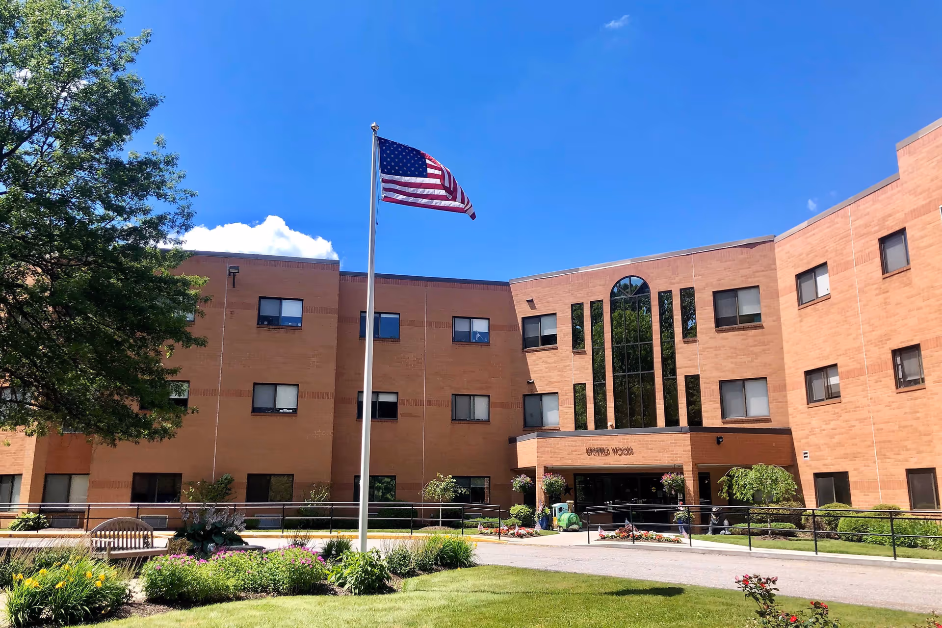 Exterior view of a three-story brick senior living facility building with multiple windows, an American flag on a flagpole in front, a well-maintained garden with flowers and shrubs, and a tree on the left side under a clear blue sky.