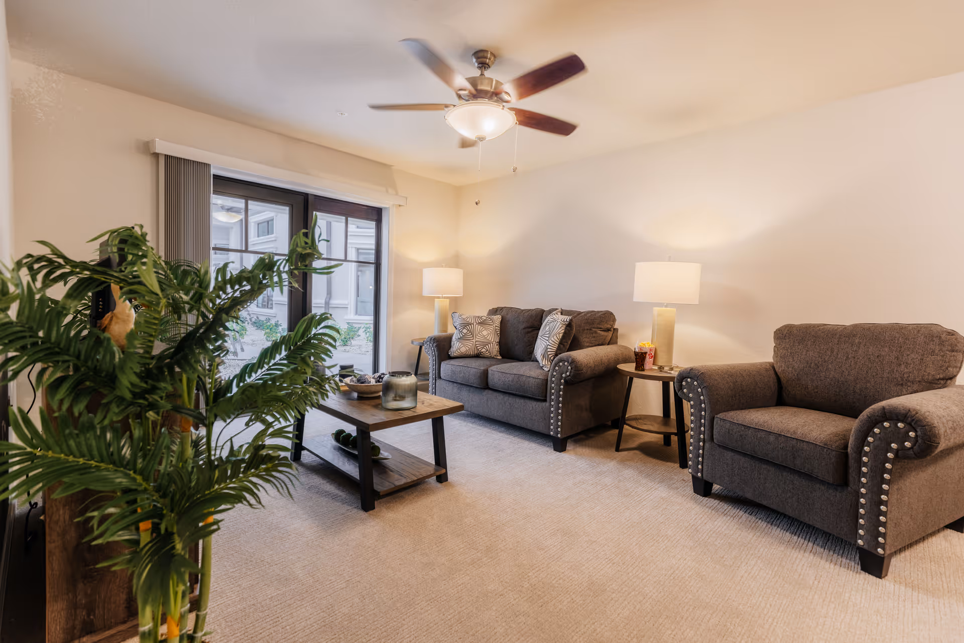 A cozy living room with a gray loveseat and matching armchair featuring nailhead trim. A wooden coffee table with decorative items sits in the center on a beige carpet. Two table lamps with white shades provide warm lighting, and a ceiling fan with a light fixture is mounted on the ceiling. A large green plant is visible in the foreground near sliding glass doors with vertical blinds, leading to an outdoor area.