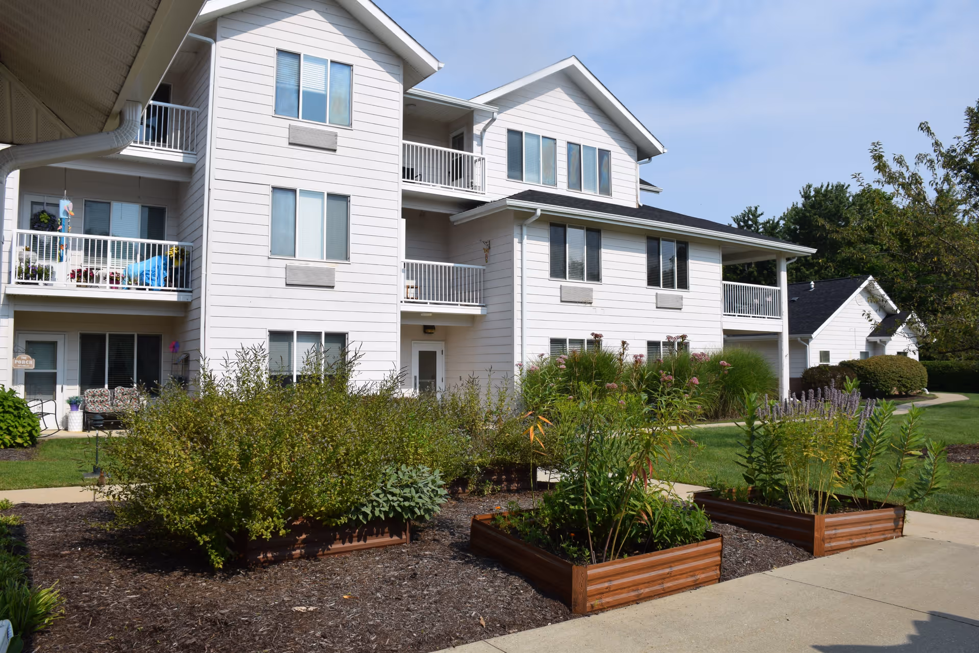 Exterior view of a multi-story white residential building with balconies, surrounded by landscaped garden beds with various plants and shrubs, under a clear blue sky.