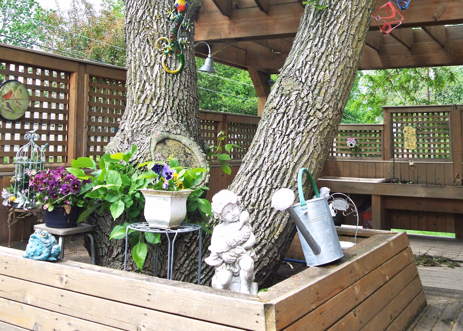 Outdoor garden area with large tree trunks surrounded by wooden fencing and lattice panels. Various potted plants and flowers are placed on metal stands and wooden surfaces. A small statue of a child holding a lamb and a metal watering can are also visible. The setting appears peaceful and well-maintained.