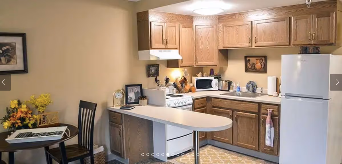 A compact kitchen area with wooden cabinets, a white refrigerator, a stove with a range hood, a microwave, and a sink. There is a small dining table with a chair and a laptop on it, along with a vase of yellow and orange flowers. The walls are beige and there are framed pictures hanging on them.