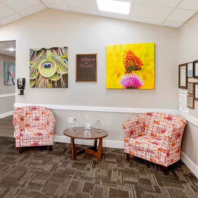 Waiting area with two patterned armchairs, a round coffee table, and colorful wall art including a butterfly canvas and a small chalkboard reading 'Welcome to Rosewood'.