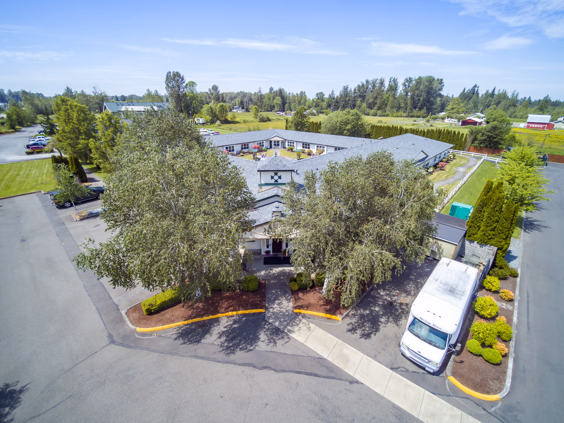 Aerial view of Heritage House Buckley Assisted Living & Memory Care facility showing a single-story building with a gray roof surrounded by trees and greenery. There is a driveway and parking area with a white van parked near the entrance. The surrounding area includes open fields and additional buildings in the distance under a clear blue sky.