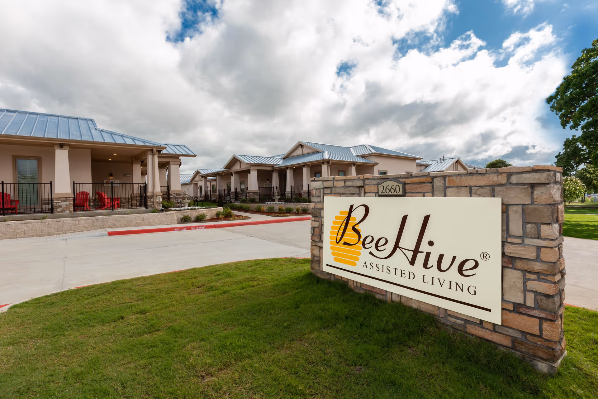 Exterior view of BeeHive Assisted Living facility showing a stone sign with the facility name and several single-story buildings with covered porches and red chairs under a partly cloudy sky.
