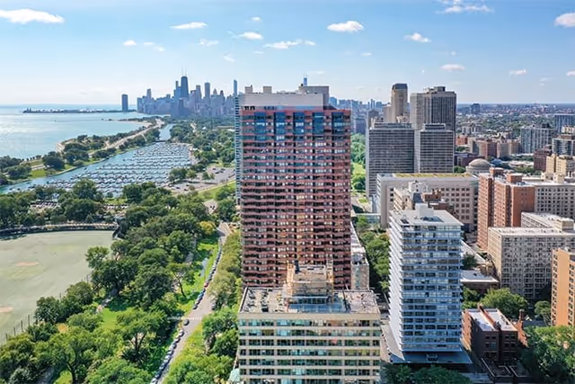Aerial view of a tall residential building surrounded by other buildings and green park areas, with a large body of water and a city skyline in the background under a blue sky with scattered clouds.