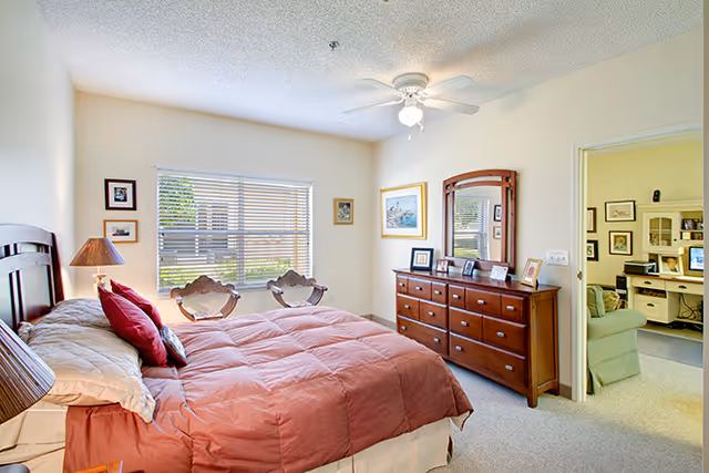Bright furnished bedroom with a bed, dresser and mirror, ceiling fan, window with blinds, and two chairs by the window.