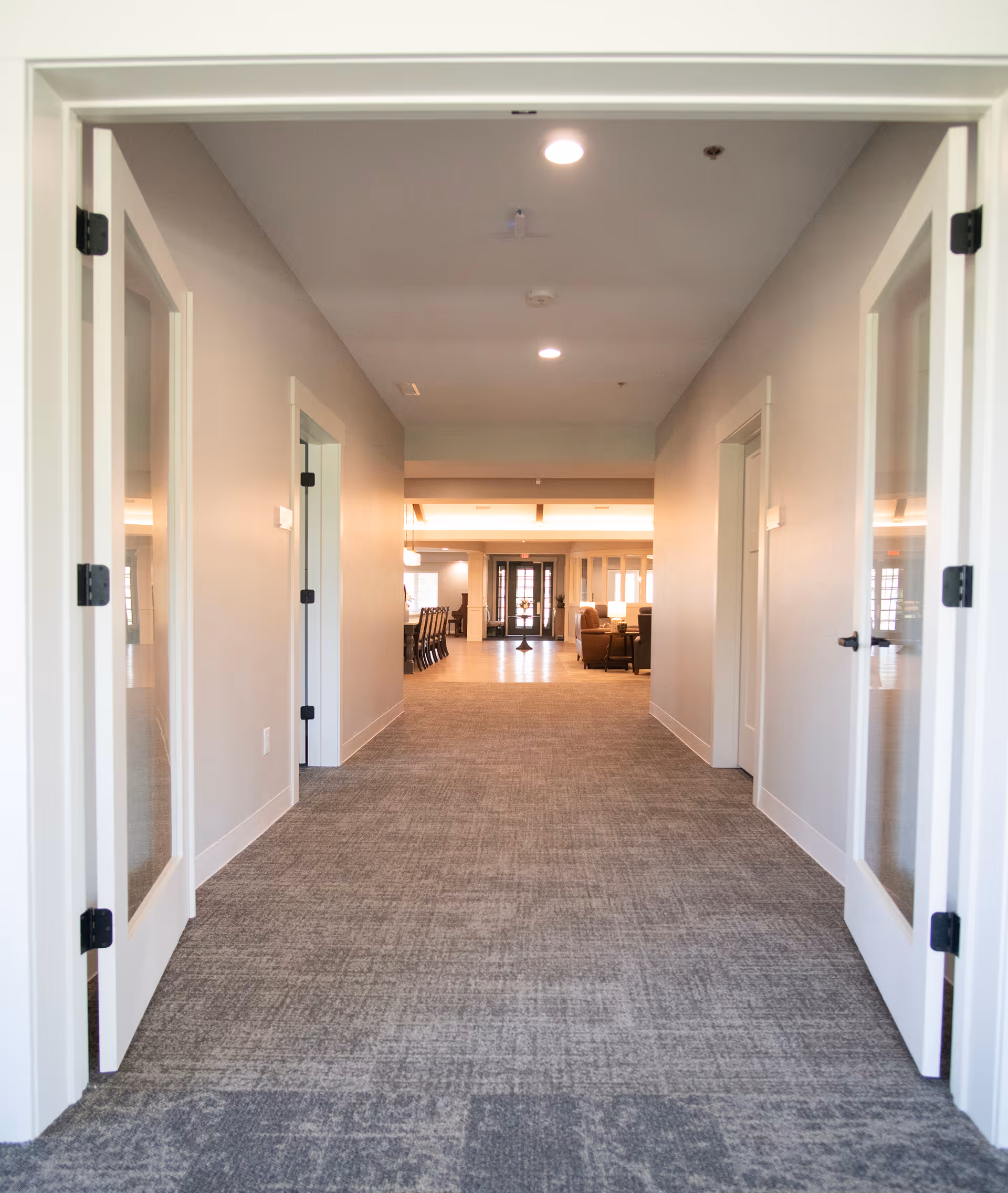 View down a carpeted hallway with open double glass doors on both sides, leading to a well-lit common area with chairs and tables in the distance.