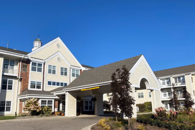 Exterior view of a senior living facility building with beige siding and brick accents under a clear blue sky. The building features multiple windows, a covered entrance with a peaked roof, and landscaped bushes and trees around the driveway.
