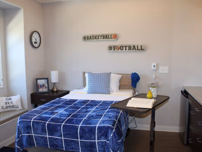 A bedroom in a senior living facility with a single bed covered by a blue checkered blanket and a decorative pillow. On the wall above the bed are two signs reading '#BASKETBALL' and '#FOOTBALL'. To the left of the bed is a nightstand with a lamp and a framed photo. A clock is mounted on the wall above a window seat with a cushion that says 'FIND YOUR HAPPINESS'. To the right of the bed is an adjustable overbed table with an open book and a small decorative item. The room has light-colored walls and dark wood flooring.