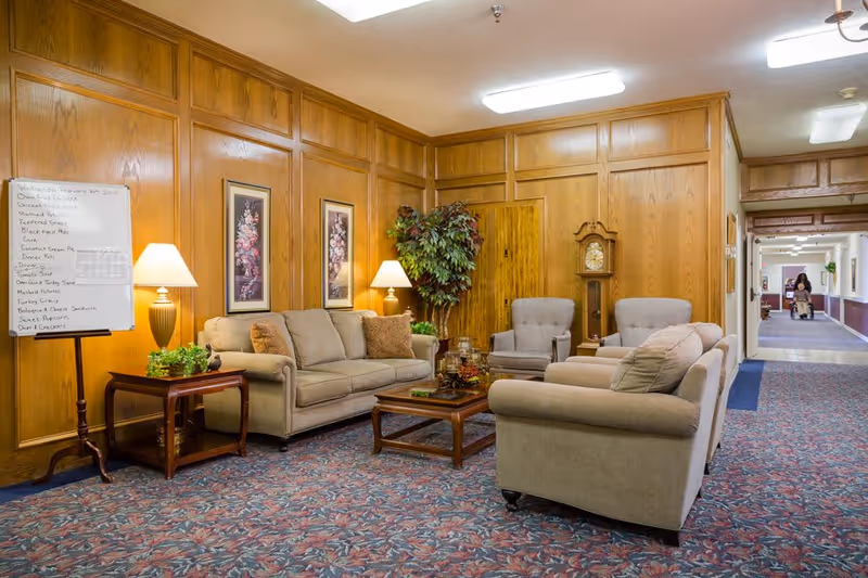 A cozy senior living facility lounge area with beige sofas and armchairs arranged around a wooden coffee table. The walls are wood-paneled with two floral paintings and a tall grandfather clock. A potted plant and two table lamps add warmth to the space. A whiteboard with a menu is visible on the left, and a hallway extends to the right with a person in the distance.