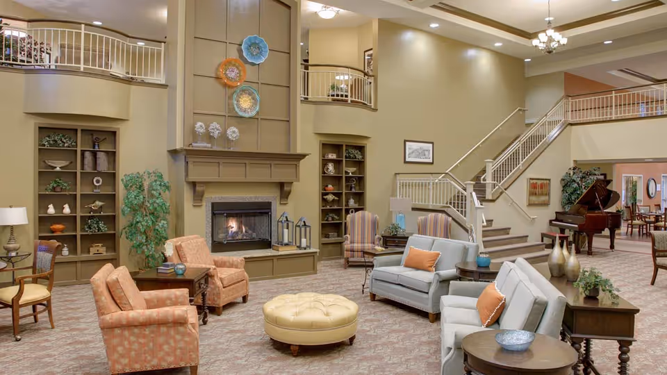 A spacious and well-lit living room area in a senior living facility featuring comfortable seating including armchairs and sofas arranged around a central round ottoman. The room has a fireplace with decorative plates mounted above it, built-in shelves with various decor items, and a grand piano near a staircase leading to an upper level. The walls are painted in warm beige tones, and the space includes side tables with lamps and plants, creating a cozy and inviting atmosphere.