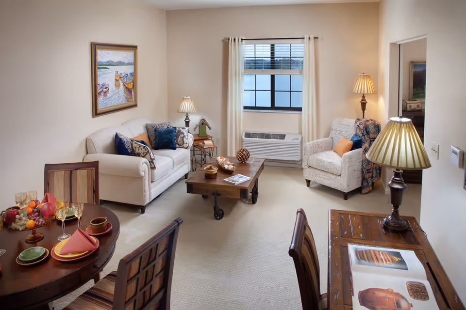 A cozy living room with a beige sofa and an armchair, both adorned with decorative pillows. A wooden coffee table with wheels sits in the center, holding a decorative bowl and a magazine. There are two table lamps with pleated shades, one on a side table next to the sofa and another on a wooden desk with an open book. A window with white curtains and a wall-mounted air conditioning unit is visible. A round dining table with four chairs is partially visible, set with plates, glasses, and napkins. A framed painting of boats on water hangs on the wall above the sofa.