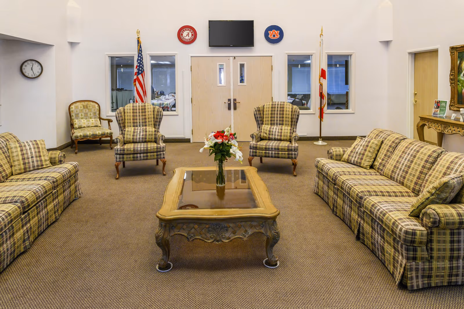 A living room area with plaid upholstered sofas and armchairs arranged around a wooden coffee table with a glass top and a vase of flowers. The room has beige carpeting, white walls, and a clock on the left wall. Two flags flank a set of double doors with a TV mounted above them. There are two small windows on either side of the doors and a decorative table with a mirror on the right side.