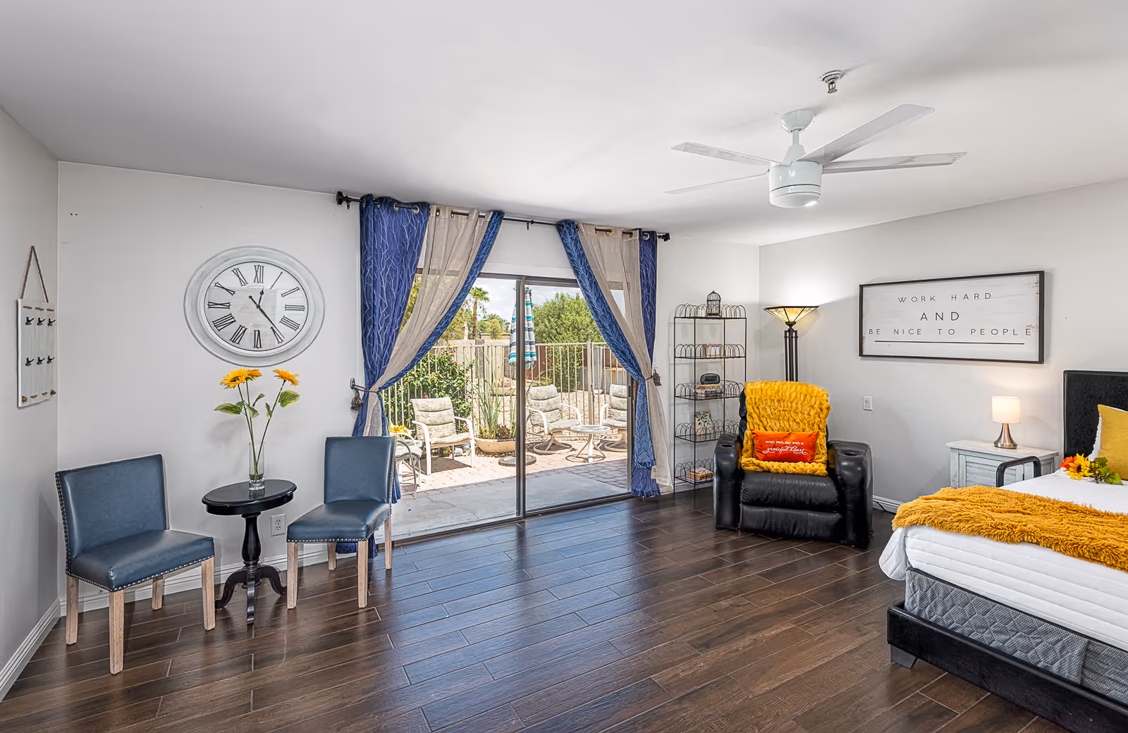 A bright and spacious bedroom with dark wooden flooring, a bed with white and yellow bedding, a black recliner chair with a yellow throw blanket, two blue chairs with a small round table between them, a large wall clock, and a sliding glass door leading to an outdoor patio with chairs and greenery.