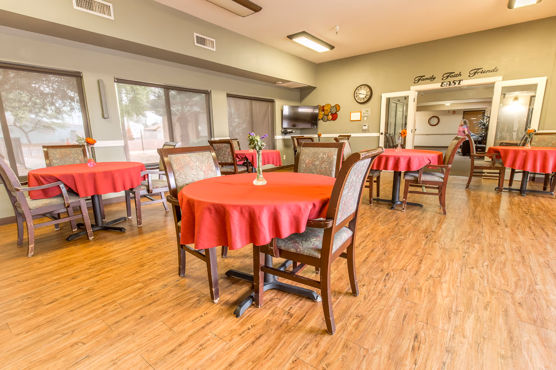 A dining room with several round tables covered with red tablecloths, each table surrounded by wooden chairs with patterned cushions. Small flower vases are placed on each table. The room has large windows with shades, wood flooring, and a wall-mounted TV. A clock and decorative wall art are visible on the far wall, along with double doors leading to another room.
