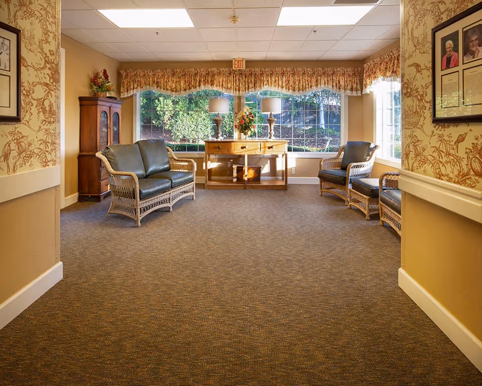 A cozy sitting area in a senior living facility with wicker furniture including a loveseat and two chairs with dark cushions. There is a wooden console table with two lamps and a flower arrangement in front of large windows with floral valances. The walls have patterned wallpaper and framed pictures, and the carpet is brown.