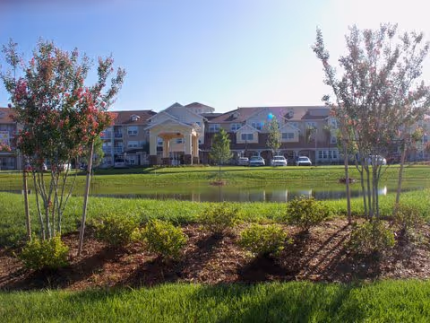 View of a large retirement living facility building with a covered entrance, surrounded by green lawns, small trees, and a pond in the foreground under a clear blue sky.