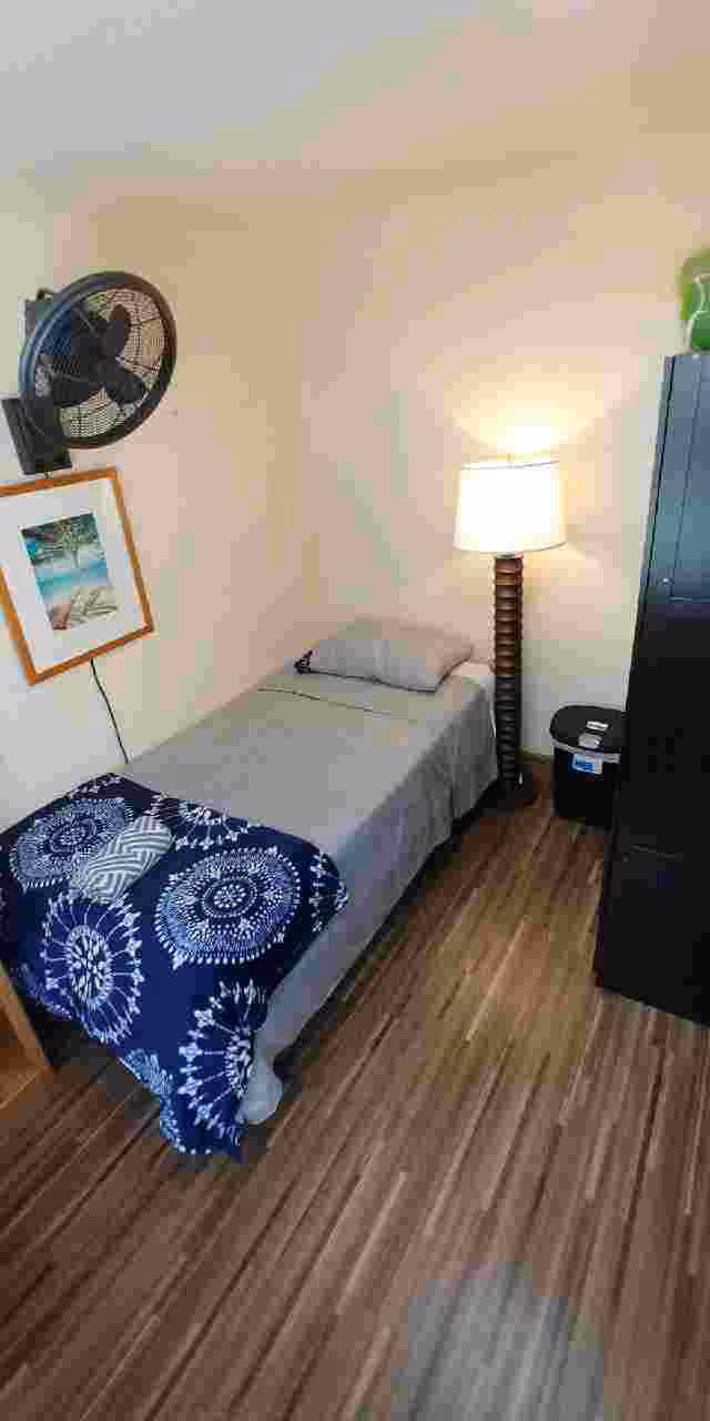 A small bedroom with a single bed covered in gray sheets and a blue patterned blanket. There is a pillow on the bed, a standing lamp beside it, a black cabinet, a trash bin, a wall-mounted fan, and a framed picture on the wall. The floor has wood-like flooring.