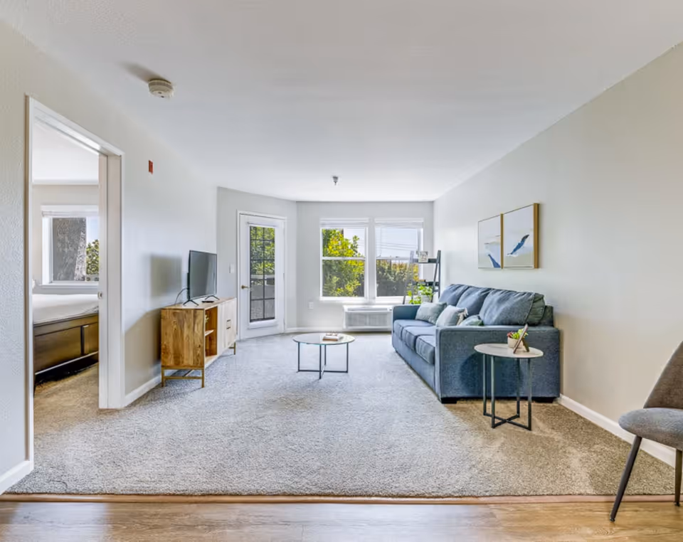 Bright furnished living room with a blue sofa, round coffee table, TV on a wooden console, and large windows and door letting in natural light.